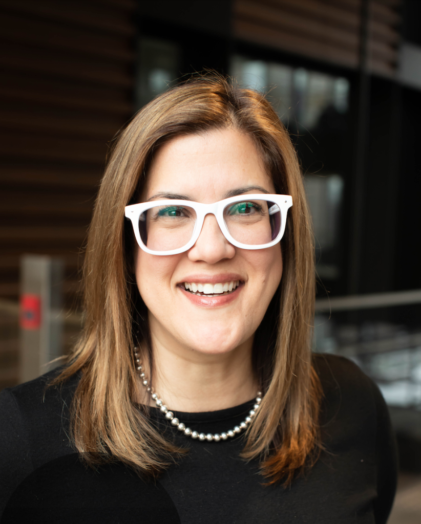 smiling woman with medium length hair and white glasses wearing a black top and a necklace
